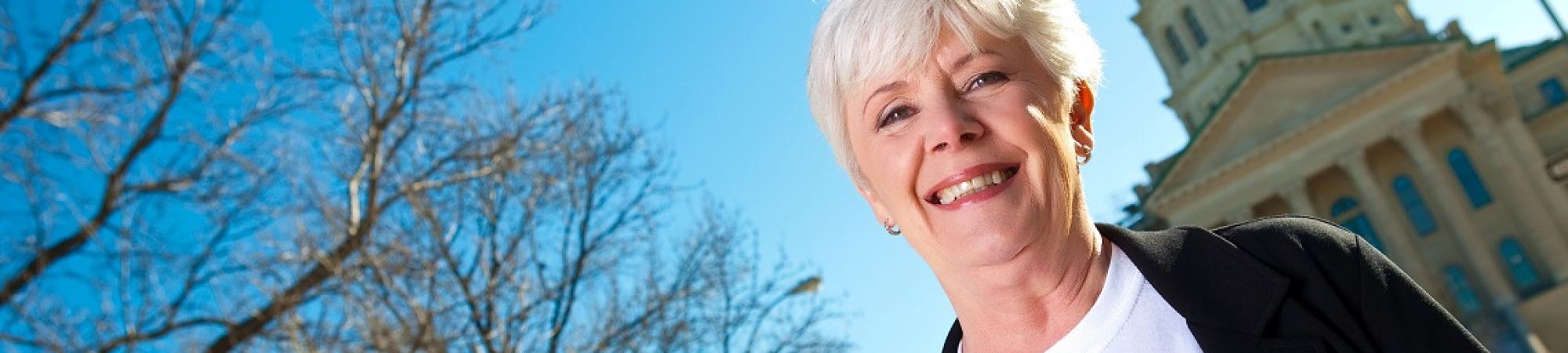 A white-haired woman wearing a white LIVE UNITED shirt under a black cardigan stands in front of the Kansas capitol building on a clear day under a blue sky. There are trees in the background with no leaves.