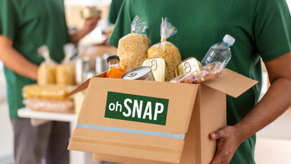 A volunteer in a green shirt carrying a box of food with a green label that says oh SNAP
